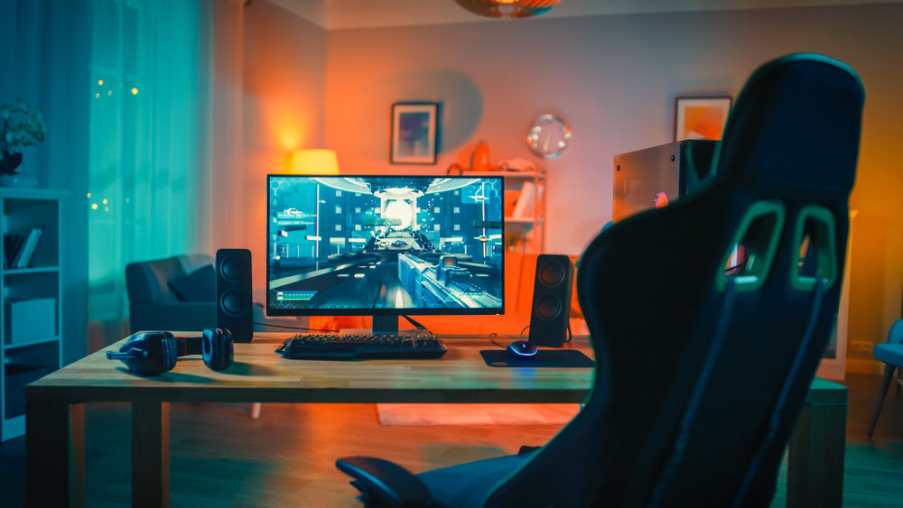 A computer with speakers and headphones showing a first-person shooter video game on the screen, and a large empty chair